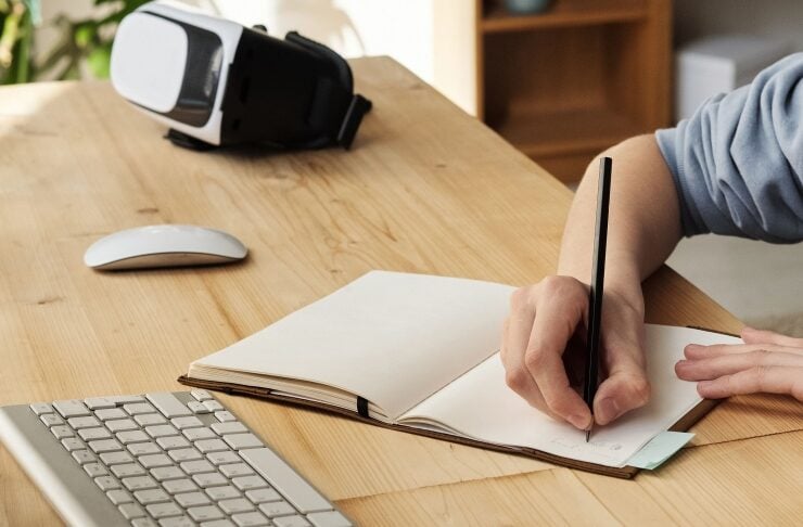 Person writes in an open notebook on a wooden desk, with a wireless keyboard, mouse, and a VR headset nearby.