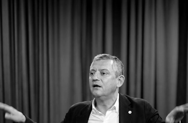 Man in a suit speaks at a panel, arms extended, with a glass of water on the table in front of him (black and white).