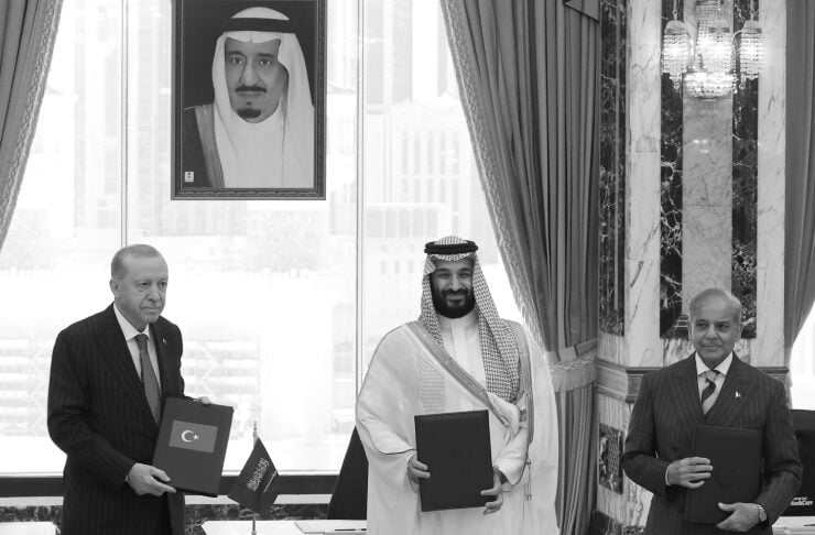 Three men in suits stand with folders during a formal signing ceremony in a grand room, a large portrait on the wall behind them.