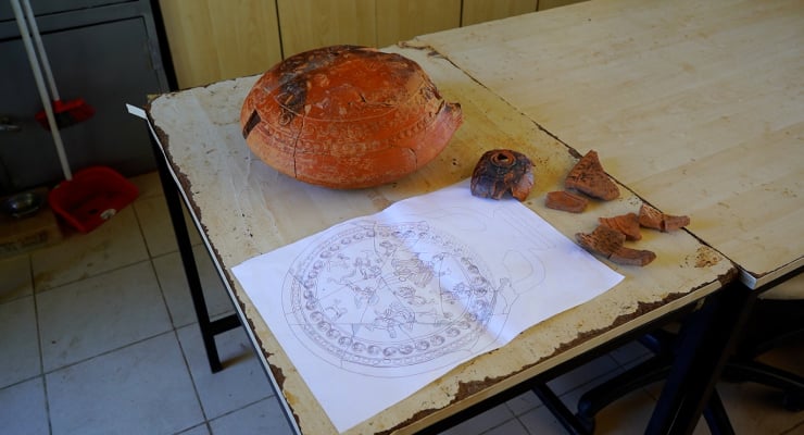 Pottery reconstruction study: a cracked orange bowl, several shard fragments, and a printed circular design on paper on a workshop table.