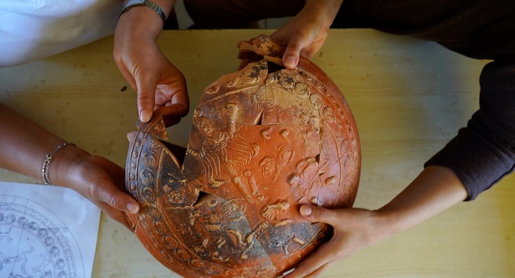 Two people hold a cracked, carved pumpkin-shaped ceramic pot with pieces being removed on a table surface, showing intricate relief design.