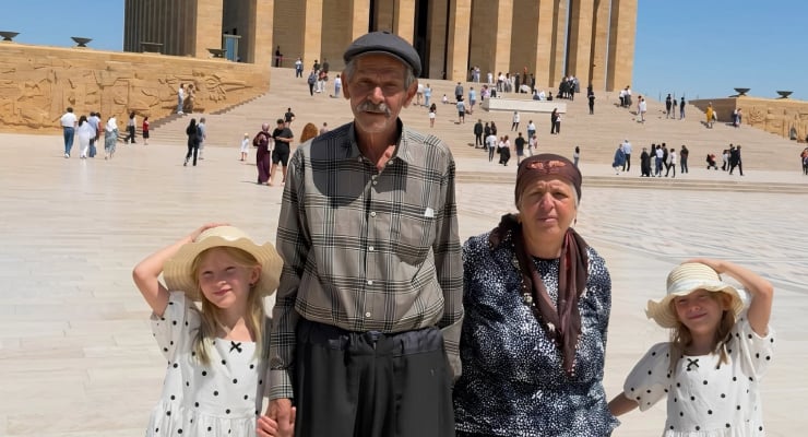 A man in a plaid shirt and cap stands with an older woman and two young girls in sun hats on the wide steps of a large monument, with visitors in the background.