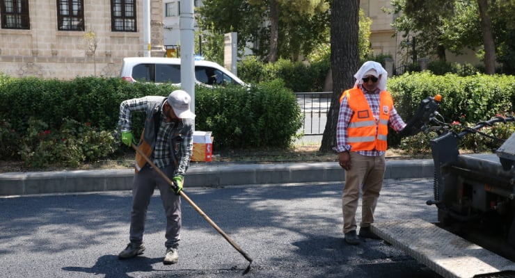 Two construction workers paving a street: one spreads asphalt with a rake while the other watches beside a paving machine, both wearing safety gear.
