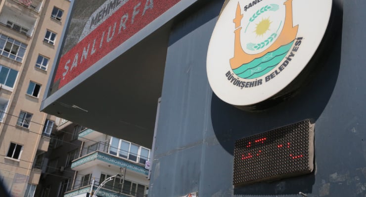 Exterior of a municipal building with the circular Şanlıurfa Büyükşehir Belediyesi logo on a blue wall and a red LED display beneath, with apartment buildings in the background.