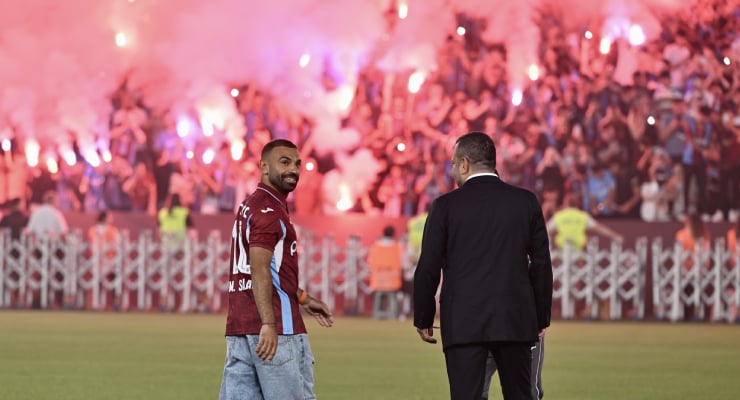 Soccer player in a maroon jersey on the field smiling at the camera as a man in a suit walks nearby, with cheering fans and red flares in the background.