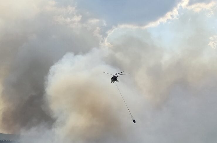 Firefighting helicopter dropping a water bucket through smoky sky over a wildfire zone.
