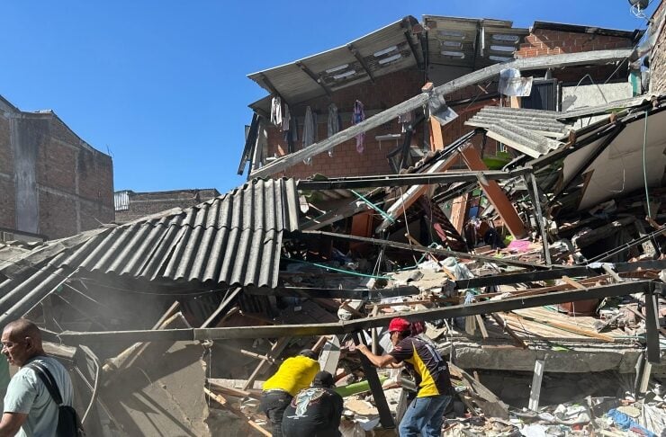 Rescue workers in yellow shirts search through rubble of a collapsed brick building under a clear blue sky.