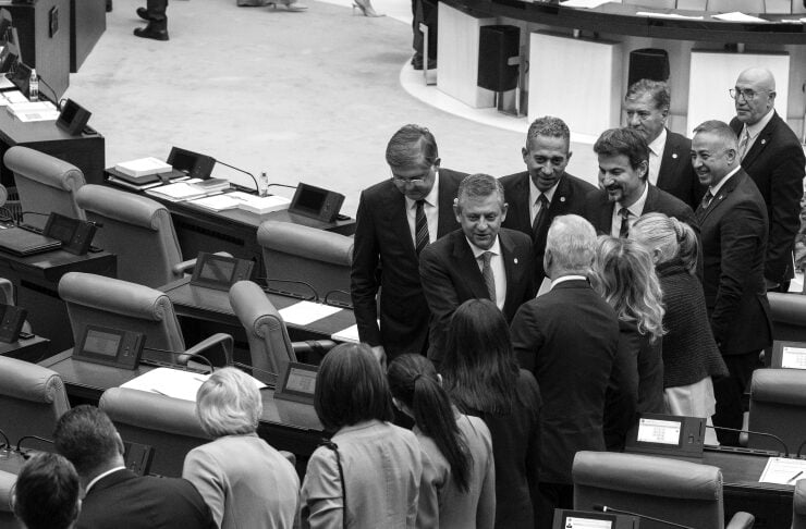 Group of formally dressed officials standing in a legislative chamber around desks with electronic devices.