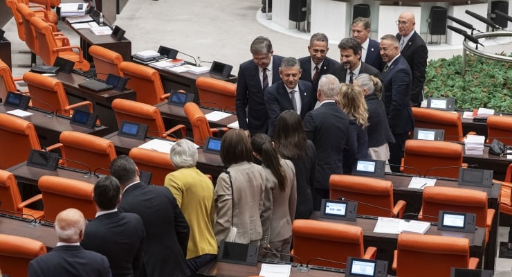 Group of suited professionals gathered and conversing in a conference chamber with orange seating and desks with tablets on them. | Note: desks appear with devices on them, suggesting a formal meeting room setting.
