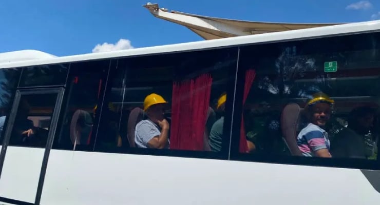People wearing yellow hard hats seated on a white tour bus with red curtains, sunny day outside the windows.