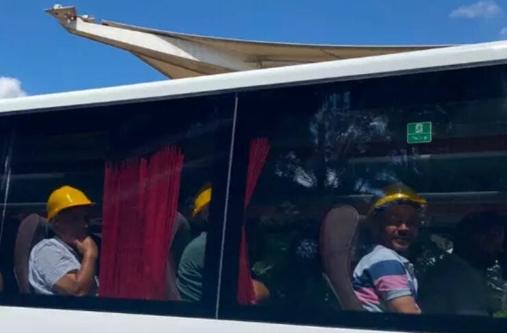 Passengers wearing yellow hard hats seated inside a white bus with red curtains, sunlit day outside.