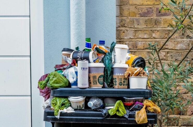Overflowing black trash bin with assorted litter and takeaway cups piled on top, against a brick wall and a white door.