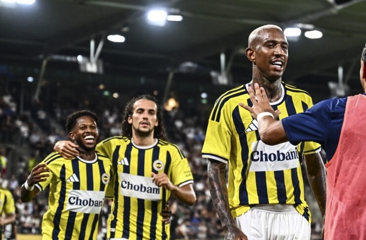 Football players in yellow and blue striped kits celebrate a goal on the pitch with a cheering crowd in the background on a night game.