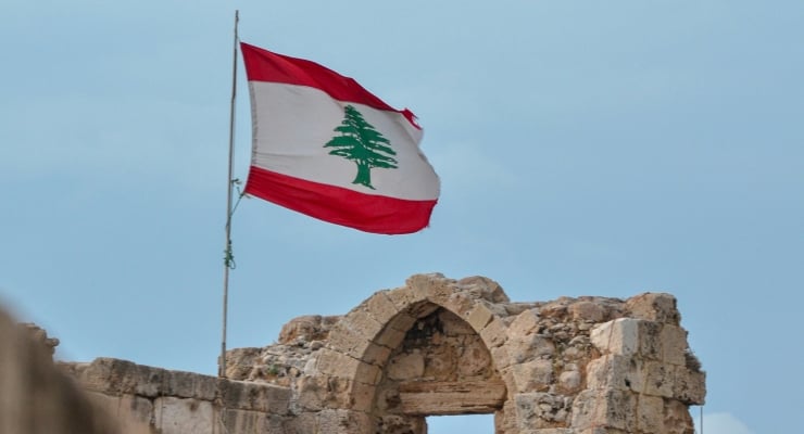 Lebanese flag waving atop a flagpole over ancient stone ruins with an arched doorway against a blue sky.