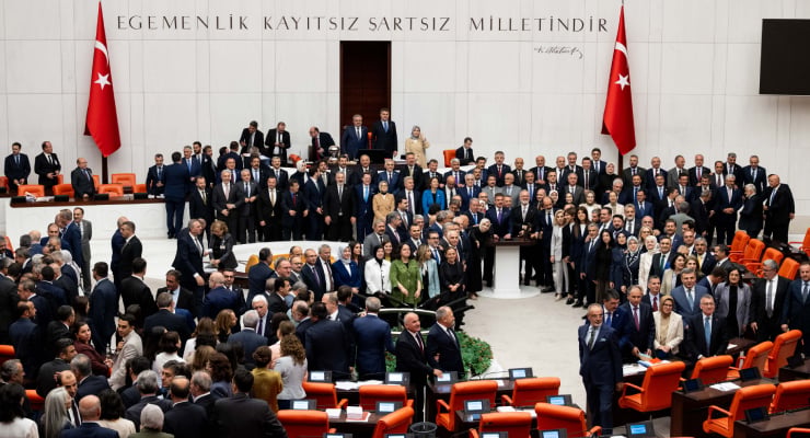 Group photo of Turkish parliament members gathered in a white marble chamber, with red seats and Turkish flags, beneath the inscription 'Egemenlik kayıtsız şartsız milletindir'.