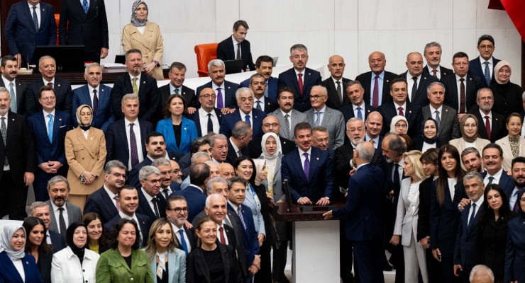 Large group of politicians and officials in formal attire gathered around a central podium as a speaker addresses the crowd in a ceremonial setting.