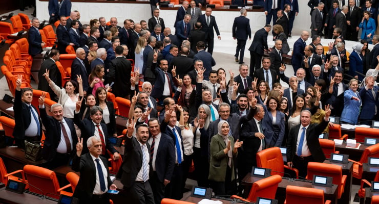 A crowded parliament chamber with politicians standing and waving from orange-red seats.