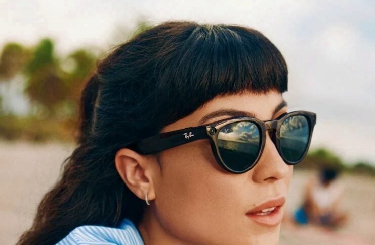 Woman at the beach wearing black Ray‑Ban sunglasses and a light blue striped shirt, looking off to the side.