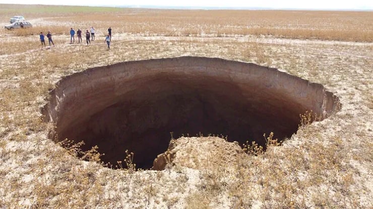 Large circular sinkhole in dry landscape with a group of people and a white vehicle in the distance.