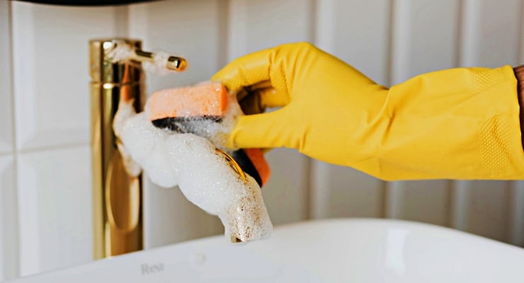 Person wearing yellow gloves scrubbing a faucet with a soapy sponge over a sink.