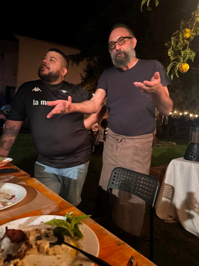 Two men stand by a table with plates of food at a nighttime outdoor gathering, one gesturing with his hands.