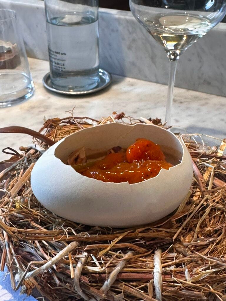 Eggshell half filled with orange sauce resting in a straw nest on a marble table, with glassware in the background.