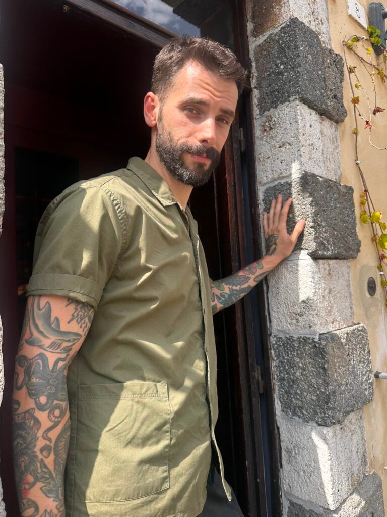 Bearded man in a green shirt leans at a rustic doorway, hand on a rough stone wall, tattoos visible on his arm.