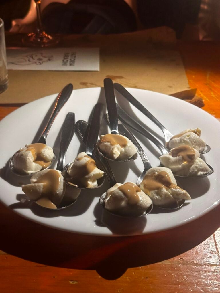 Plate of small oysters placed on metal spoons, each topped with a creamy brown sauce, on a white plate at a dimly lit table.