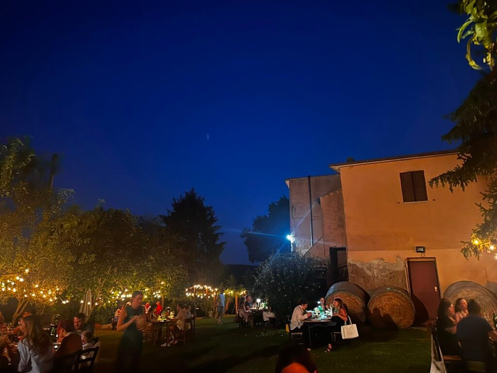 Outdoor evening dining scene with string lights, groups of people at tables under trees by a peach-colored building.