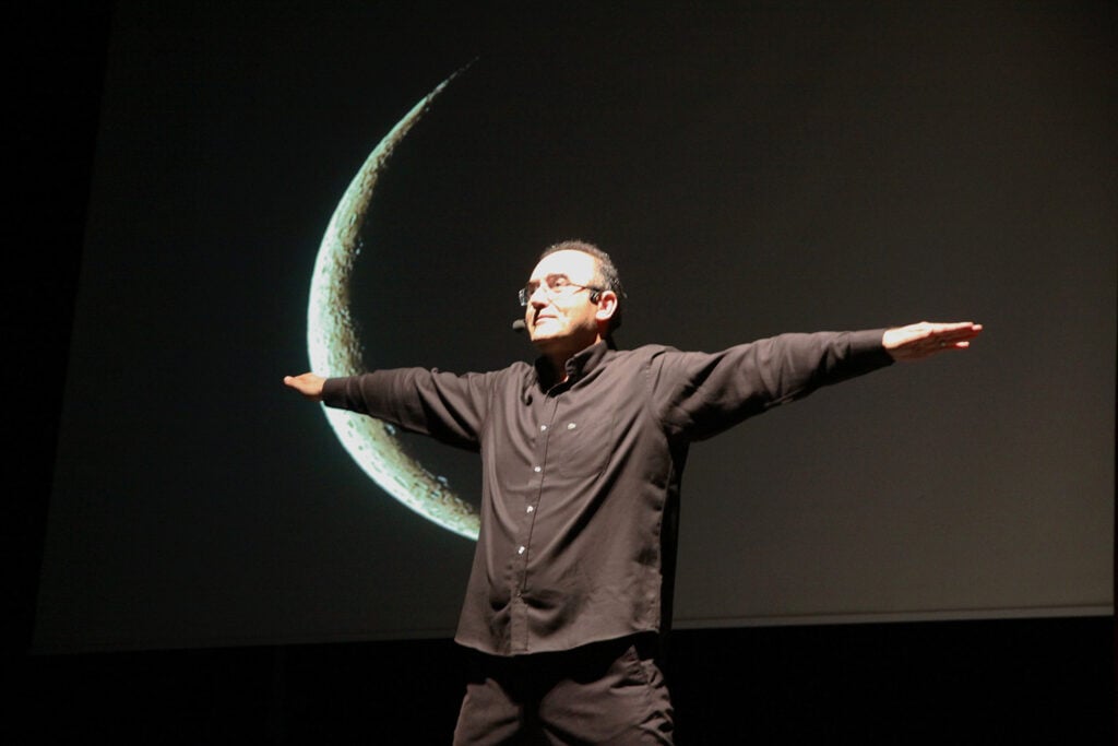 Man on a dark stage with arms outstretched, wearing a black shirt, in front of a large crescent moon projection behind him.