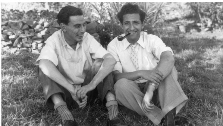 Two men sit on grass outdoors, smiling and talking; one holds a pipe in his mouth.