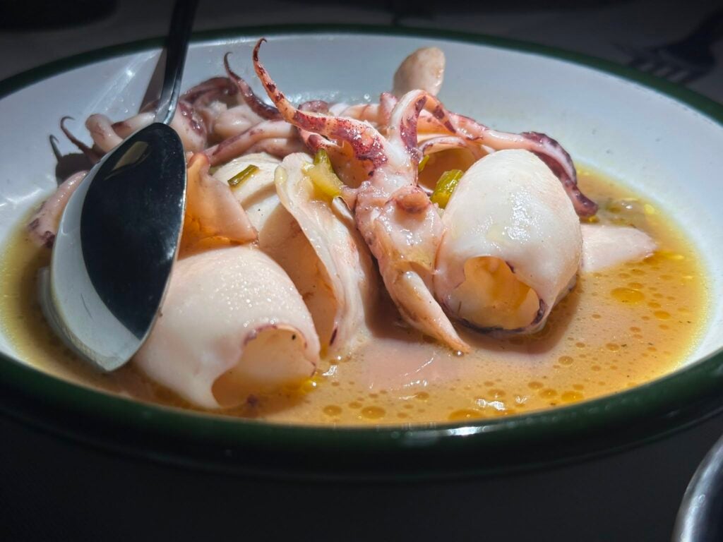 Bowl of seafood soup with squid tentacles and white fish in orange broth, spoon resting on the edge of the bowl.