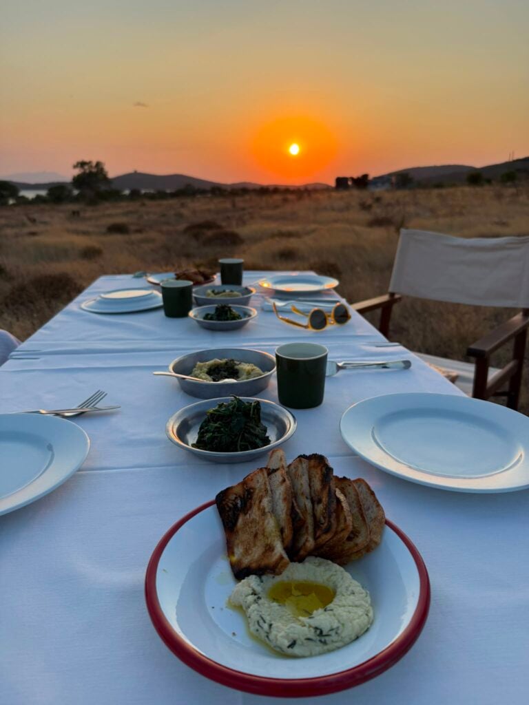 Outdoor dinner setup at sunset: a long white table with bowls of greens, bread, and plates glowing in warm light. A chair is at the right, and a bright sun sits low on the horizon.