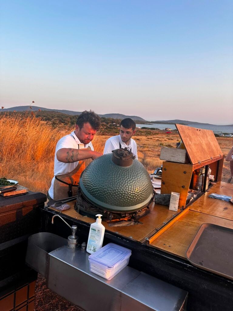 Two men in white shirts and aprons cook at a large green kamado grill outdoors near a coastal hillside.