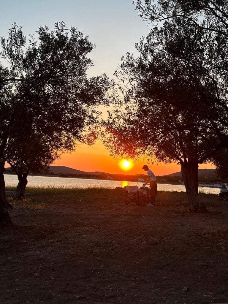 Sunset over a lakeshore framed by trees, with a person tending a small outdoor table and chairs nearby.