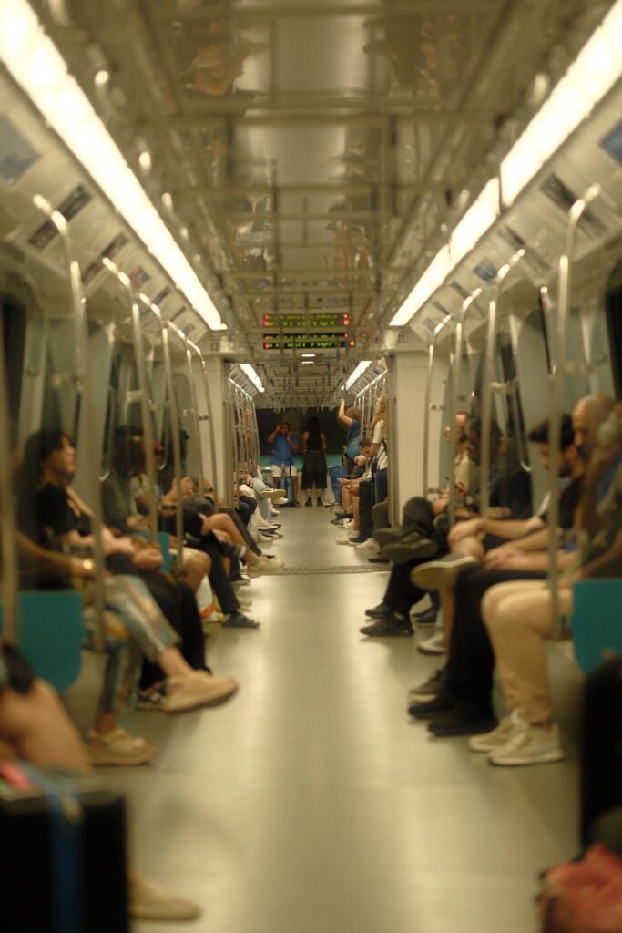 Inside a crowded subway car, passengers seated along both sides and a few standing in the aisle, lit by overhead lights